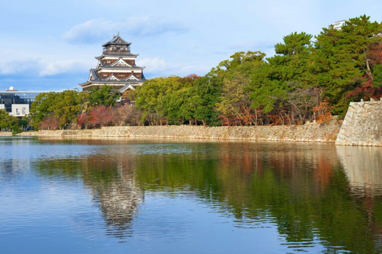Traditional architecture of Hiroshima Castle beautifully reflected in the serene waters and lush green trees.